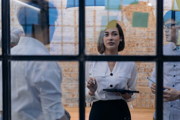 20s Asian company female manager brainstorming in the meeting room while having sticky notes on glass in office. She's holding a tablet while working. Cooperation and teamwork concept