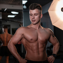 Strong young guy bodybuilder with a muscular naked torso poses on camera in the hall