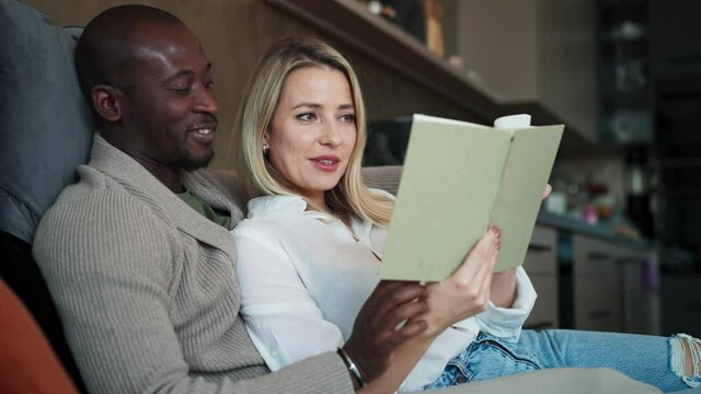 Mid adult biracial couple in love sitting and reading book together at home.