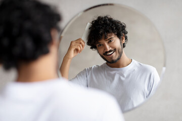 Male haircare. Positive indian bearded guy brushing hair with hairbrush, looking at his reflection in mirror in bathroom