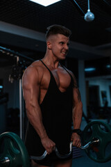 Fototapeta na wymiar Handsome young emotional strong athlete man with a muscular body and a black T-shirt does an exercise and workout in the gym on a dark background. Healthy lifestyle
