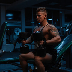 Handsome young athlete male bodybuilder trainer with muscular body and tattoo sitting and doing exercise with dumbbells in the gym
