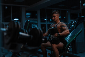 Handsome young man bodybuilder with muscular body sits with dumbbells and pumps muscles in a gym with exercise equipment. Sports lifestyle