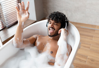 Joyful indian guy in wireless headphones enjoying music and singing while lying in hot bubbly bath...