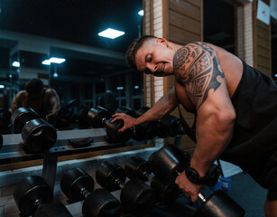 handsome sports man bodybuilder with a muscular body and a maya tattoo pumps up his biceps and exercises with dumbbell in the gym