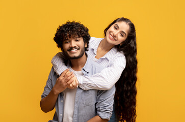 Lovely indian woman hugging her husband from behind, smiling and feeling happy on yellow studio background
