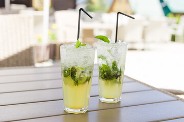 Two glasses of Lemonade or mojito cocktail with lemon and mint, cool refreshing drink or beverage, outdoor. Cold detox water with ice and straw, copy space.