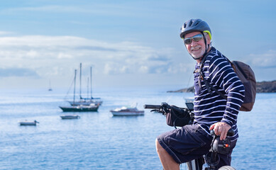 Smiling adult mature senior cyclist man wearing sunglasses, backpack and sport helmet riding in front of the sea enjoying outdoors, freedom, healthy lifestyle. Happy active elderly during retirement