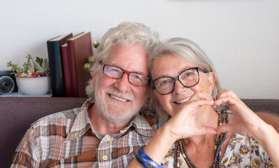 Portrait of attractive adult senior couple of grandparents making heart gesture with fingers. Elderly pensioner showing love and demonstrating sincere feelings sitting indoors, looking at camera