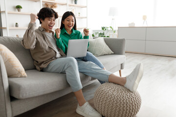 Emotional Asian couple with laptop making YES gesture, celebrating success, sitting on couch at...