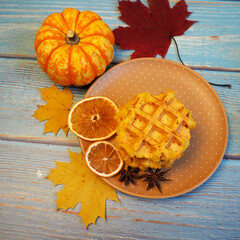 Belgian gluten- and lactose-free waffles with pumpkin stand on a brown plate with a small pumpkin on a blue table with yellow maple leaves. autumn