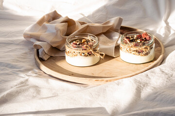 Fototapeta na wymiar Homemade granola parfait. Quick healthy breakfast greek yogurt, granola with dried berries and nuts in glass jar on wooden tray in white bedding in morning sunlight. Copy space, selective focus.