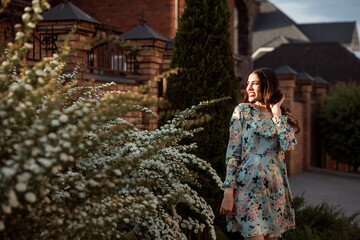 Beautiful woman in romantic dress with flowers is posing next to blooming bush, sunbeam on her face