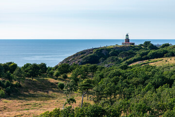 Kullaberg fyr, old lighthouse on the cliffs of Kullaberg naturreservat on the Swedish west coast. Lighthouse on the rocks viewing out into the baltic sea. Warm summer, high cliffs, deap ocean. Trees - obrazy, fototapety, plakaty