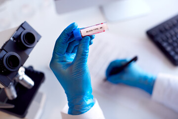 Close Up Of Lab Worker Conducting Research Using Microscope Holding Blood Sample Labelled Type AB