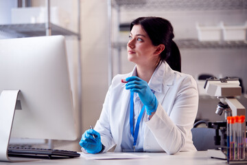 Female Lab Worker Wearing PPE Face Shield Recording Blood Test Results On Computer