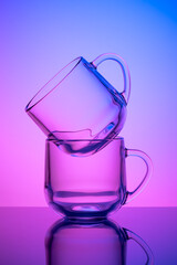Two empty tea cups on a pink and blue neon background. Drinking glass. Dark silhouette, night illuminated effect. Glassware. Transparent tableware, dishes.