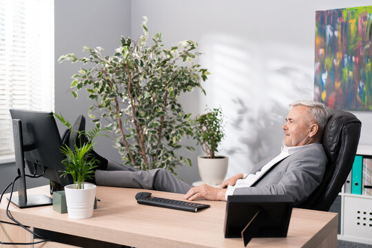 A relaxed smiling elderly company boss sits in a comfortable chair in a relaxed position with feet on the desk, a man with gray hair in suit relaxes in office after hard work in front of the computer