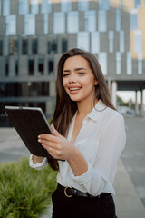 Fototapeta premium pretty woman in white shirt stands in front of glass modern business building of insurance corporation company, women is holding tablet in hand, browsing web, answering email, reading messages