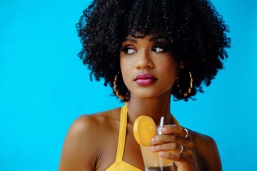 beautiful young African American woman in bright summer outfit drinking refreshing cocktail isolated on blue background