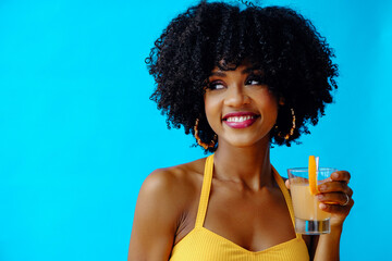 beautiful young African American woman in bright summer outfit drinking refreshing cocktail isolated on blue background