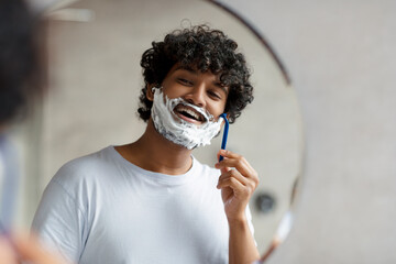 Portrait of indian man shaving near mirror in bathroom, guy with shave foam on face using razor for...