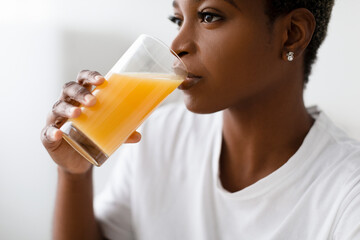 Cropped thirsty young pretty black lady in white t-shirt drinks fresh orange juice from glass...