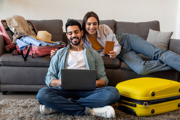 Excited couple using laptop to book tickets and hotel online, preparing for vacations and packing suitcases