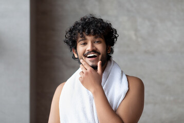 Portrait of happy bearded indian man with towel posing in bathroom and touching chin, ready to take bath