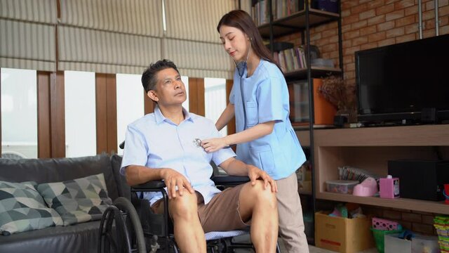 Young Asian female physical therapist standing using a stethoscope put on chest of a middle-aged Asian male patient sitting on a wheelchair. Physical examination while doing therapy treatment at home.
