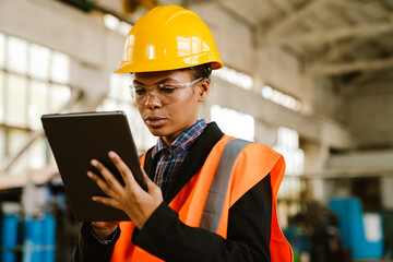 Black woman wearing helmet working with tablet computer at factory