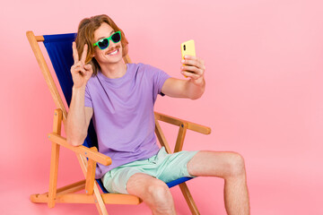 Portrait of handsome trendy cheery guy sitting in chair taking selfie showing v-sign isolated over...