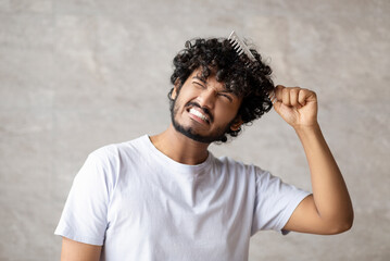 Dry and matted hair. Indian young guy in white t-shirt combining with brush and having pain from...