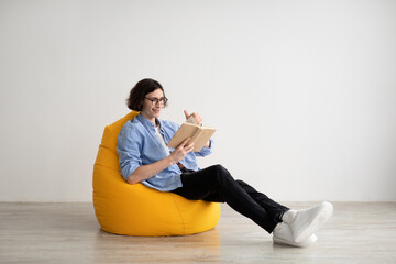 Relaxed guy reading book while sitting in comfortable beanbag chair over light wall, free space....