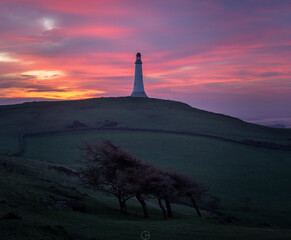 A tower on a hill with a sunrise behind under a beautiful pink sky and trees in the foreground 