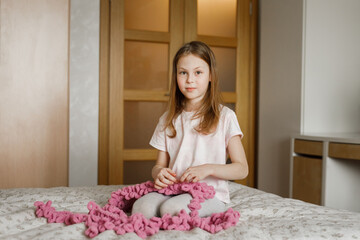 Child girl knitting from fluffy yarn sitting on bed in real bedroom interior.