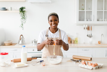 Cheerful young pretty black female in apron breaks egg, prepare baking pie or pizza
