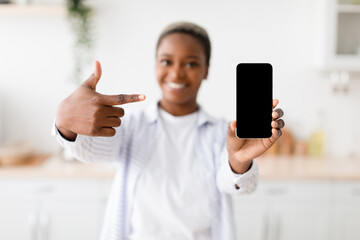 Cheerful young black woman shows on smartphone with blank screen at scandinavian kitchen interior, selective focus