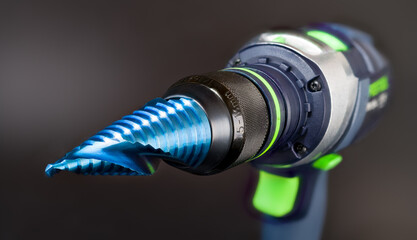 Blue nano coated cone step drill bit clamped in cordless screwdriver on gray blur background....