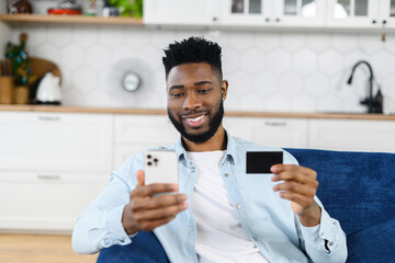 Smiling multiracial man shopping online sitting on the sofa at home, holding credit card and smartphone. Dark skinned multiracial guy making food order, purchasing online, booking and paying