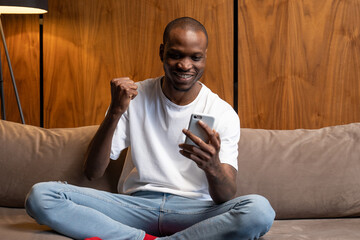 Cheerful black man using a smartphone is sitting at home on the couch, celebrating a victory, winning an online lottery, rejoicing in victory