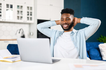 Smiling man sitting at the laptop, holding hands behind head, relaxing, looking at the screen, while working in comfortable office. Man did the job productively and got praise