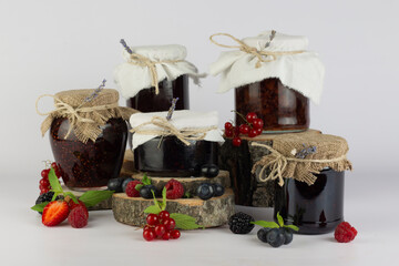 Assortment of seasonal berries jams with mint and berries. Round natural wooden podium with jars of sweet jam on white background.