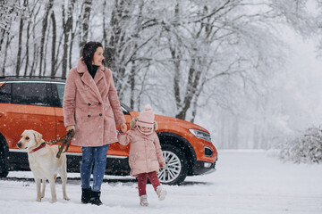 Young mother with little daughter and pet walking in a winter park by the car