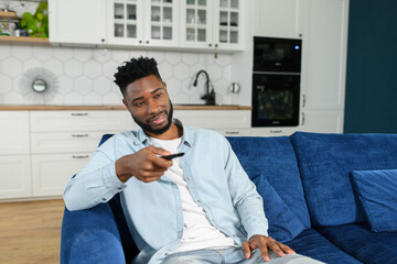 Happy young multiracial man holding tv remote control while sitting on a couch at home and relaxing at the weekend. Stock photo