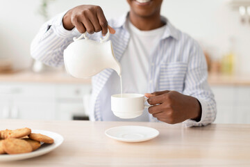 Satisfied young black woman pours milk at cup of favorite drink, sits at table with cookies in kitchen interior