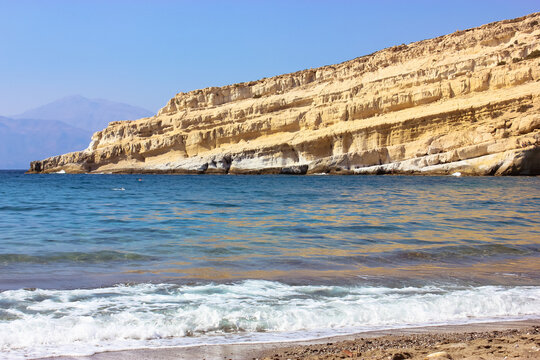 Sandy Matala beach with cliff, cliffs along a sea shore, caves against blue sky on seaside coast in Greece, Crete. Ocean waves. Resort for summer holidays without people. Mountain beach landscape.