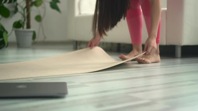 Closeup view of sportive woman spreading mat and sitting in front of laptop on floor at home spbd. Young beautiful female puts on rubber and sits down, uses computer before starts workout in light