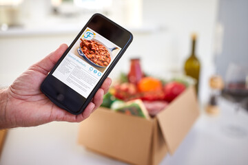 Hand In Kitchen Holding Phone With Recipe For Online Meal Food Recipe Kit Delivered To Home