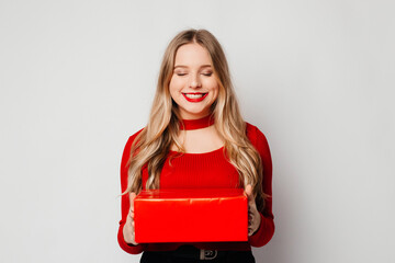 Attractive smiling blonde girl with closed eyes holding red Valentine gift box, receiving a present...
